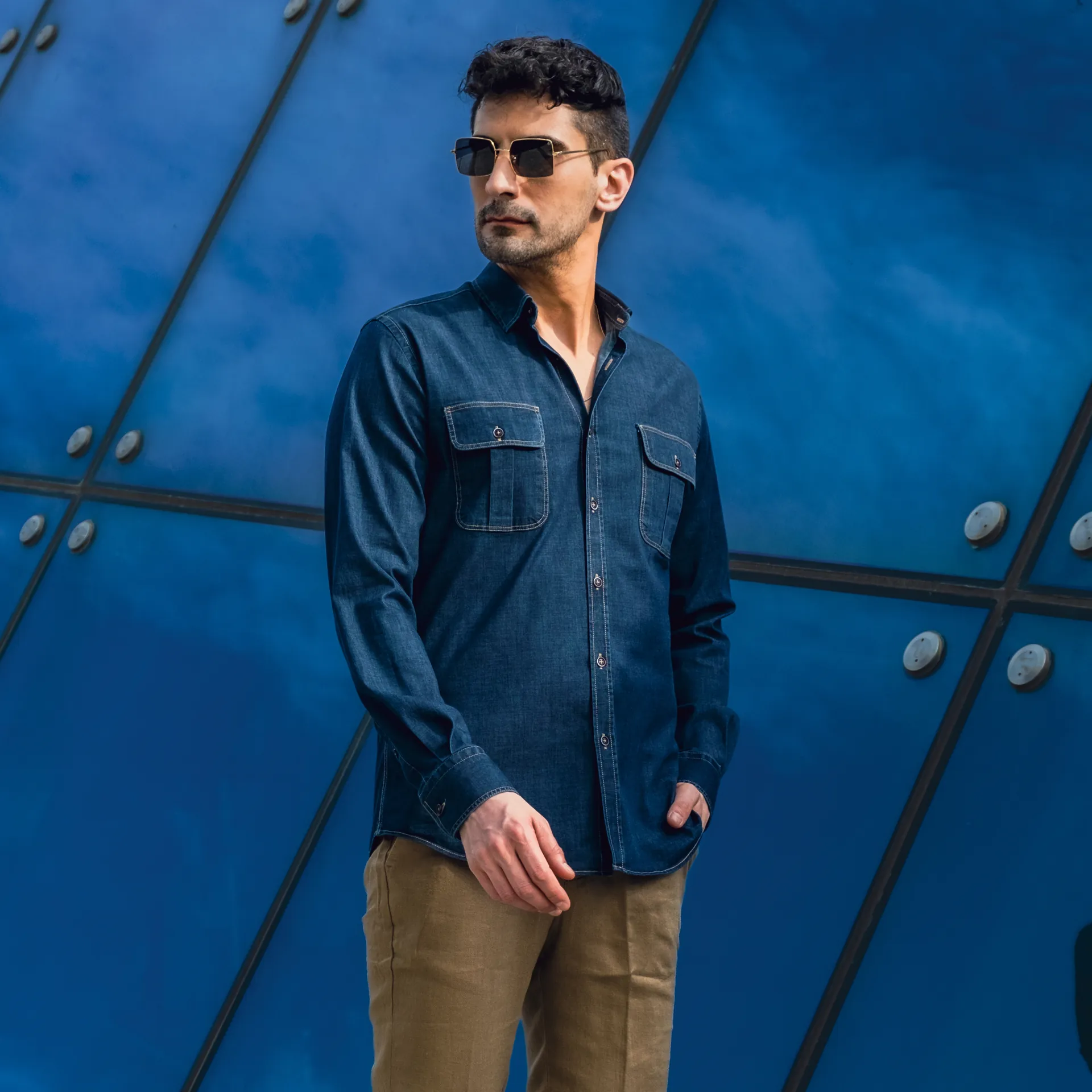 A man in a denim shirt and sunglasses against a blue glass facade.